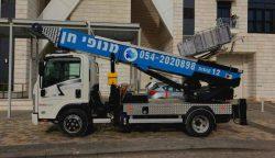 מבט מהצד של משאית חונה כחול-לבן מצוידת במעלית הניתנת להרחבה, המשמשת באופן בולט להובלת מנוף, באזור עירוני. הרכב כולל טקסט בעברית ומספר טלפון בצדו. ברקע נראית חזית בניין מודרנית עם אלמנטים מזכוכית ואבן.