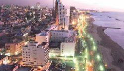 מבט אווירי של נוף עירוני חוף בשעת בין ערביים, הכולל בניינים רבי קומות מוארים וכביש הומה שלאורכו אורות עזים. הסצנה התוססת מזכירה את היעילות של חברת הובלה מומלצת, המחברת בצורה חלקה את הליבה העירונית התוססת עם קו החוף הנרחב הנמתח למרחקים.